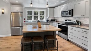 A modern kitchen with white cabinetry, a stainless steel refrigerator, a built-in microwave, and a large island with stools. - Long Sands Your All-season Getaway (York)