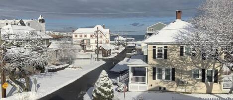 A residential street with houses, a snow-covered road, and a body of water in the background.