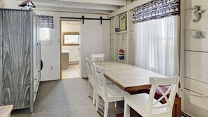 Un comedor con mesa de madera y sillas blancas, una cocina con armarios blancos y vista al exterior a través de una ventana con cortinas ligeras.