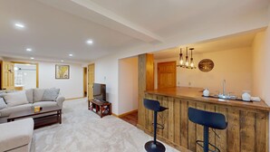 A living room with a sofa, TV, and coffee table. There's a wooden bar with stools and a kitchen area with a sink and countertop. - Beechwood Home in Mendon (Rutland)