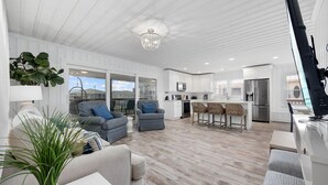 A modern living room with a large window, a dining area with a bar, and a kitchen with white cabinetry. - Summer Breeze in North Myrtle Beach (North Myrtle Beach)