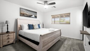 A bedroom with a wooden bed, a ceiling fan, a painting, a window with blinds, a bedside table with a lamp, and a TV mounted on the wall. - Devonshire in Scottsdale (Scottsdale)