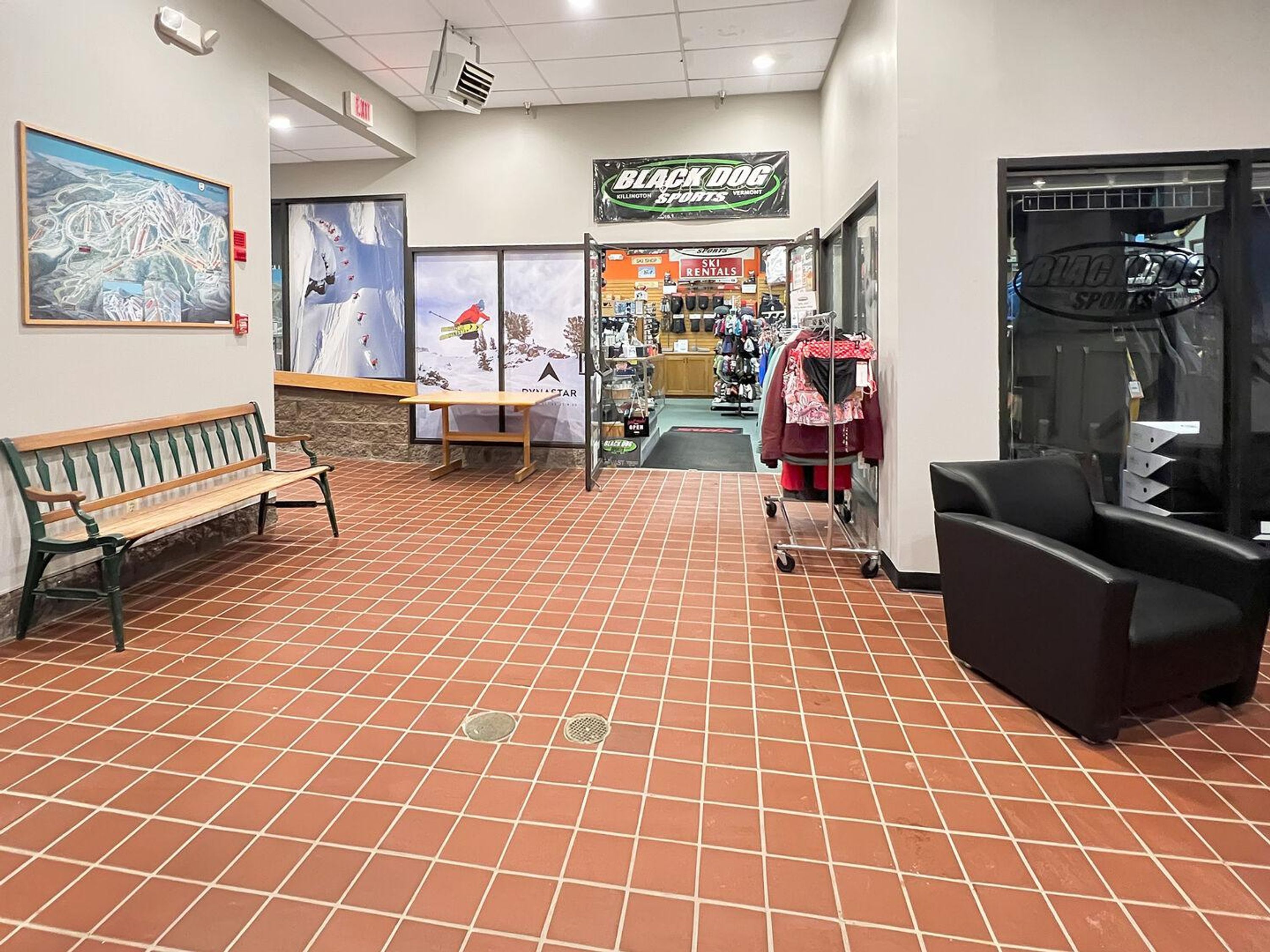 A lobby with a tiled floor, a bench, a wall-mounted sign, and a storefront with ski rental services.