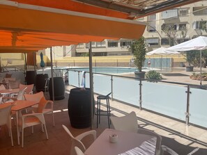 Un espacio al aire libre con mesas y sillas, una piscina al fondo y un edificio con balcones.