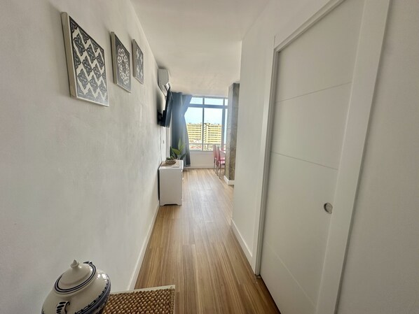Un estrecho pasillo con piso de madera, una puerta blanca y un comedor con mesa y sillas visibles a través de una ventana abierta.
