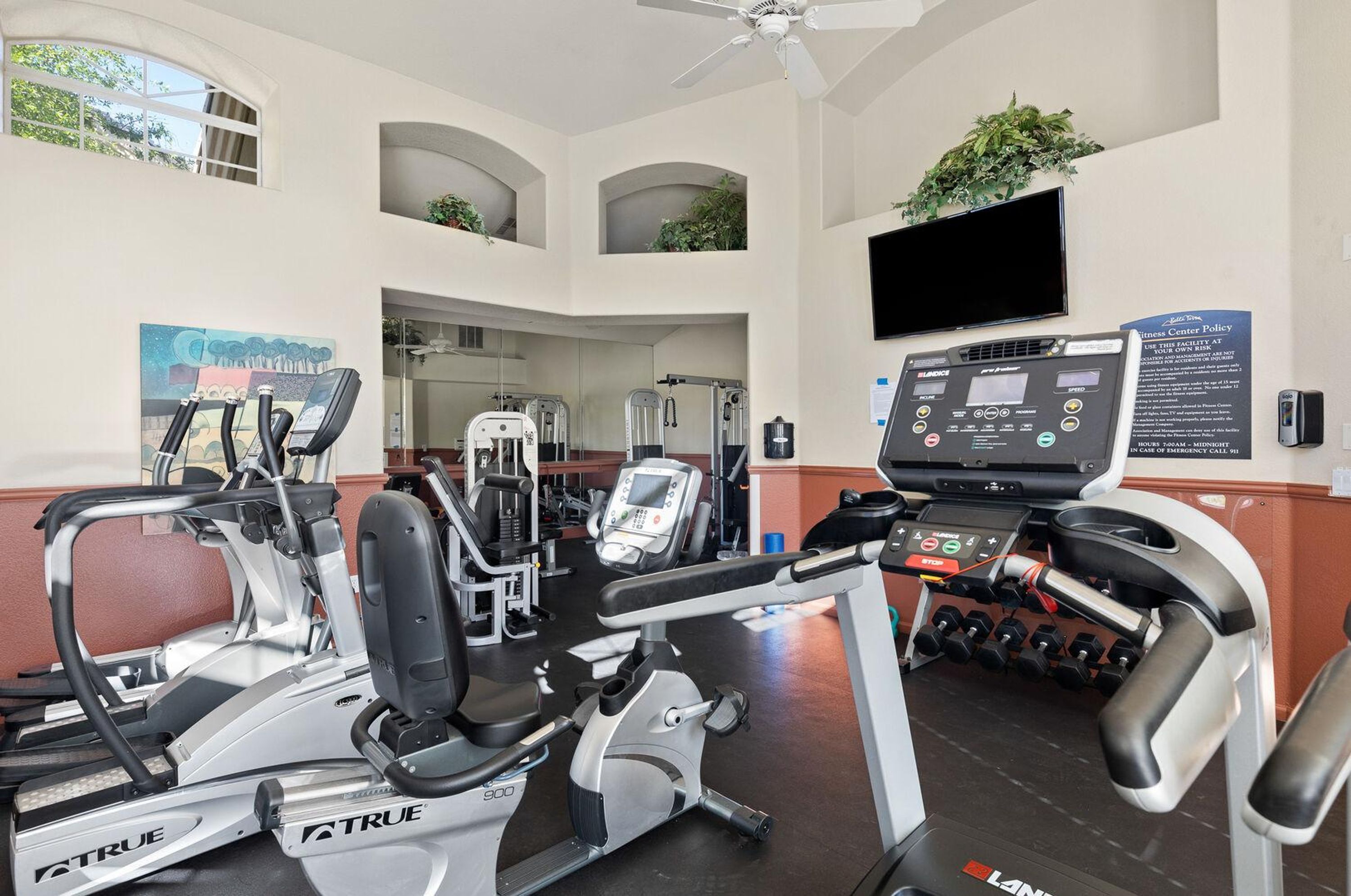 Une salle de sport équipée de tapis de course, de vélos elliptiques et d’appareils de musculation. Un téléviseur est fixé au mur, et un panneau affiche le règlement du centre de remise en forme.