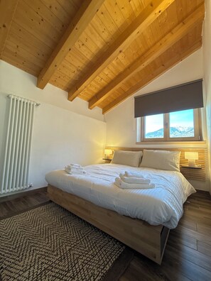 A bedroom with a wooden ceiling, a bed with white linens, a window with a view of mountains, and a radiator. - Apartment quiet area, ideal for relaxing as a couple or with family (strembo)