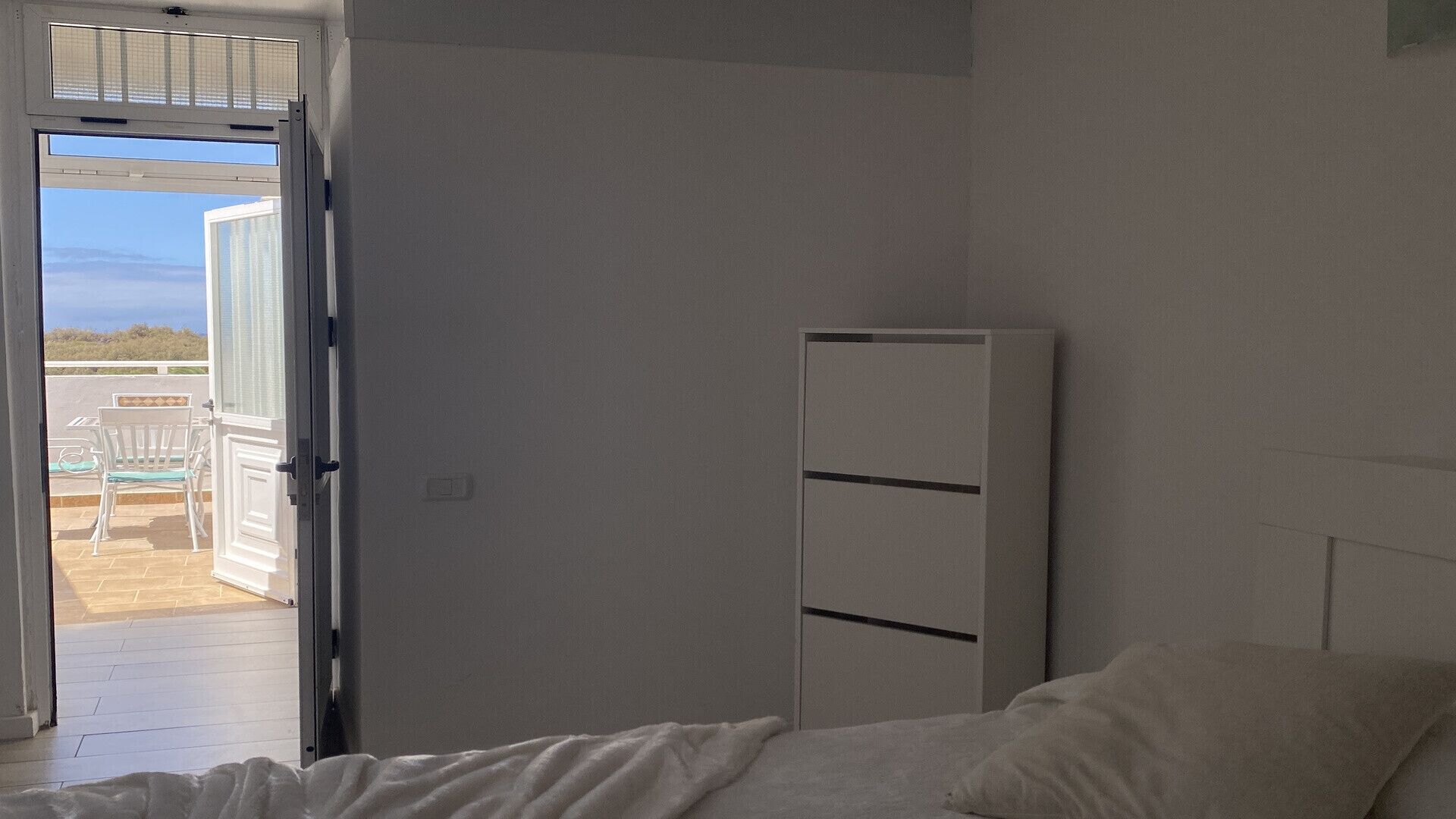 A bedroom with a bed, a white wardrobe, and a door leading to an outdoor area with a view of the sea.