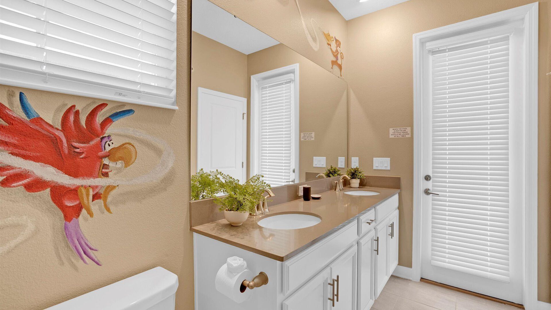 Une salle de bain avec un mur de couleur beige, un grand miroir, un lavabo avec un plan de toilette beige, des toilettes et une porte avec un cadre blanc.
