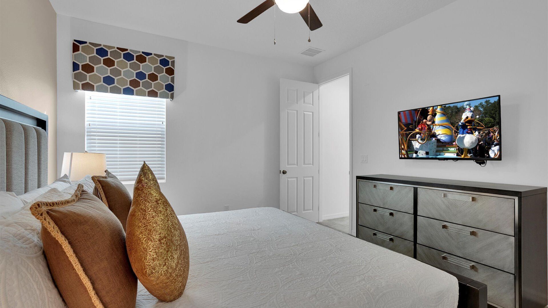 A bedroom with a bed, pillows, a dresser, a ceiling fan, a window with blinds, and a television on the wall.