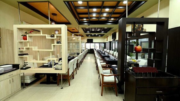 A long dining area with multiple tables and chairs, a kitchen area with cabinets and appliances, and a ceiling with recessed lighting. - HOTEL SAGAR INTERNATIONAL (Durg)