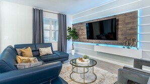 Een moderne woonkamer met een blauwe bank, een glazen salontafel en een tv aan de muur.