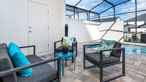 An outdoor patio area with two grey sofas, one with a blue pillow, and a small table with a cactus. A pool is visible in the background. - ADT8976 Summary:<br>Sola (Kissimmee)