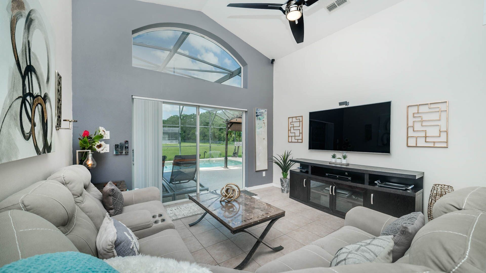 A modern living room with a large TV, comfortable seating, a glass door leading to a patio, and a ceiling fan.