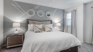 A modern bedroom with a large bed, two bedside tables with lamps, a dresser, and decorative mirrors on the wall.