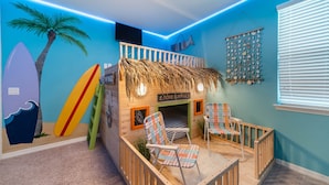 A children's play area with a beach theme, including a playhouse, surfboards, and a wall with a surfboard mural.