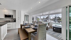 Une cuisine moderne avec des meubles blancs, un four à micro-ondes et un coin repas offrant une vue sur une piscine et des palmiers.