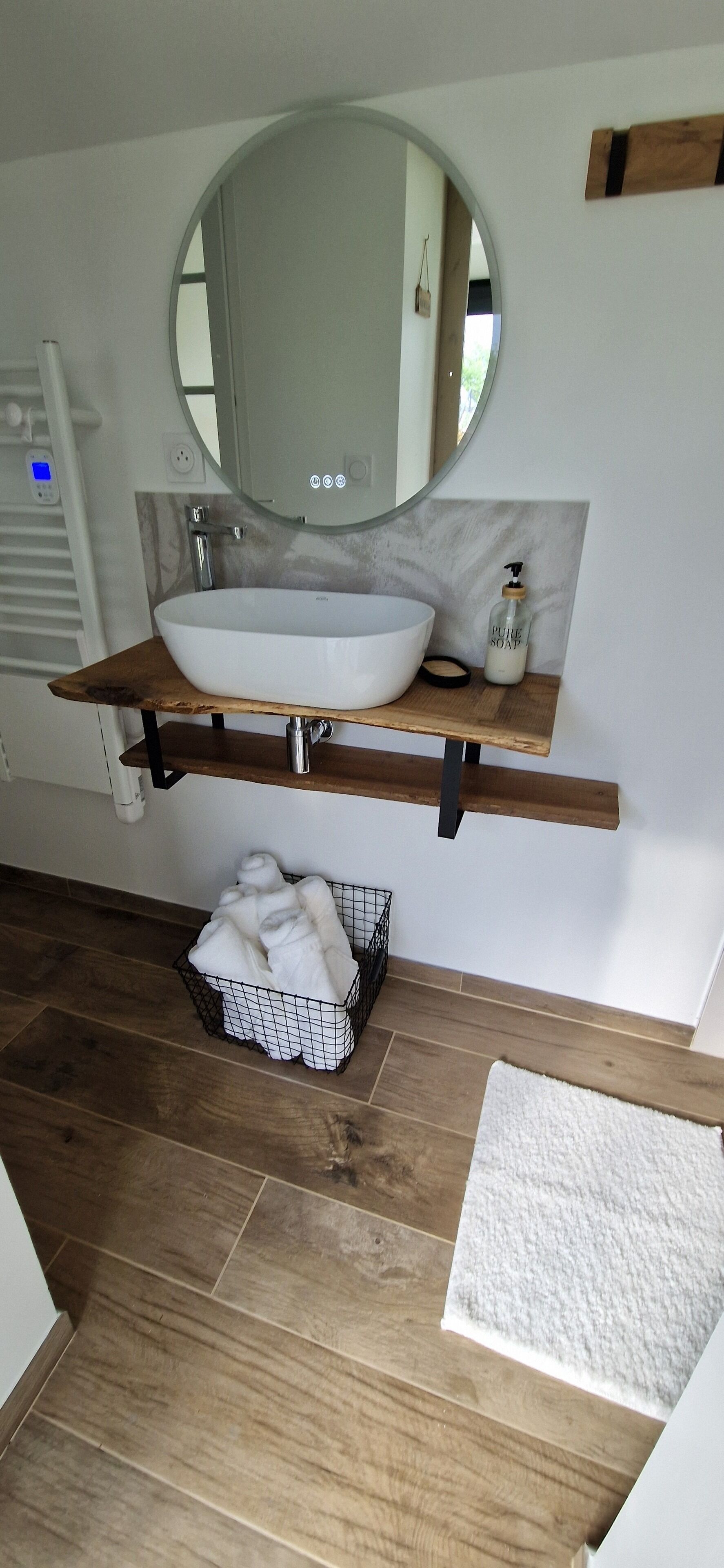 A bathroom with a white sink, a round mirror, and a wooden shelf. There is a white towel basket on the floor.