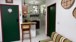 Een woonkamer met een groene deur, een gestreept bankje, een houten stoel, en een keuken in de achtergrond, evenals een plafondlamp.