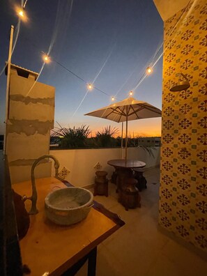 A rooftop terrace with a table, chairs, a large umbrella, and string lights.