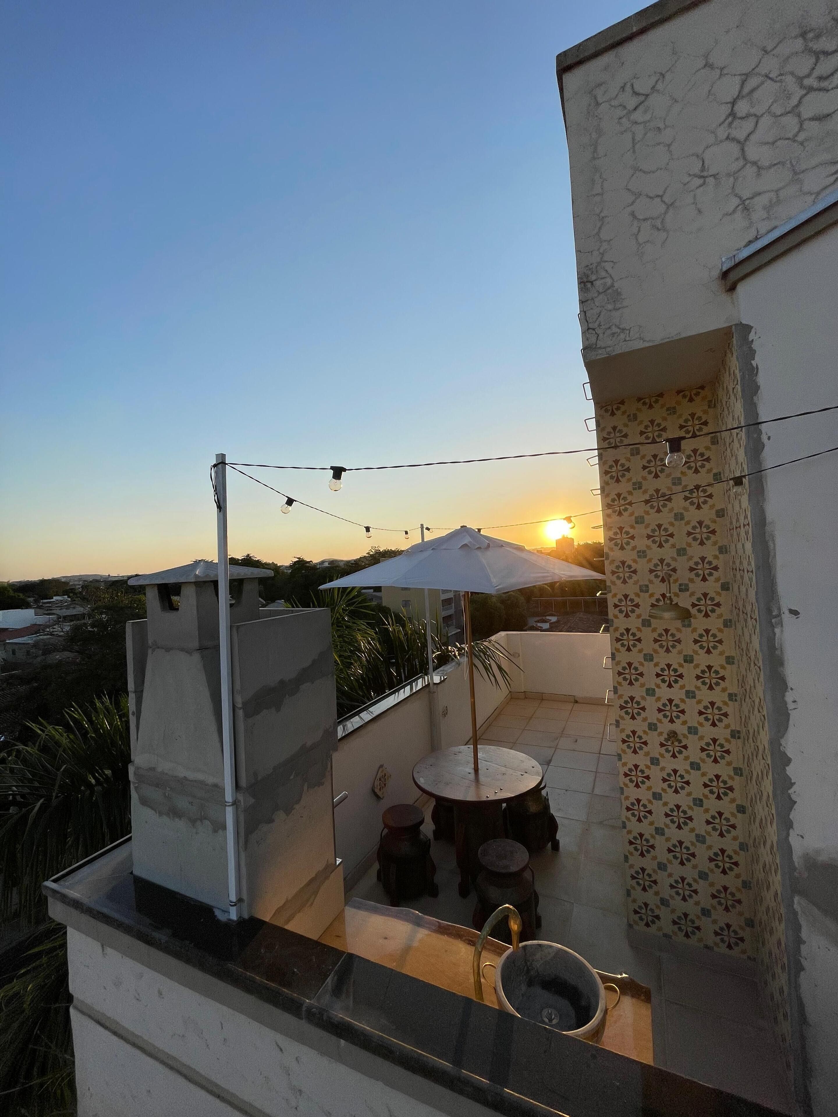 Una terraza en la azotea con mesa y sillas, un pequeño lavamanos y vistas al atardecer.