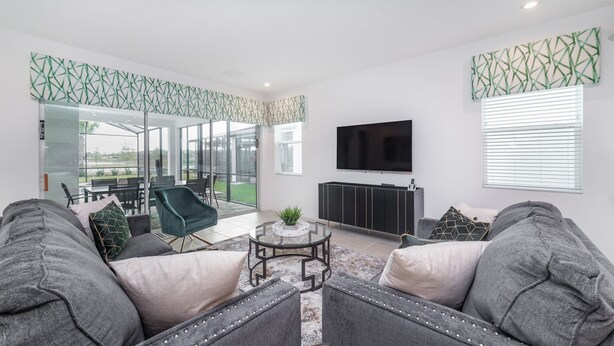 A modern living room with a large TV, grey sofas, and a glass door leading to an outdoor area.