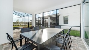 A modern outdoor dining area with a stainless steel table and chairs, large glass windows, and a view of a swimming pool and greenery.