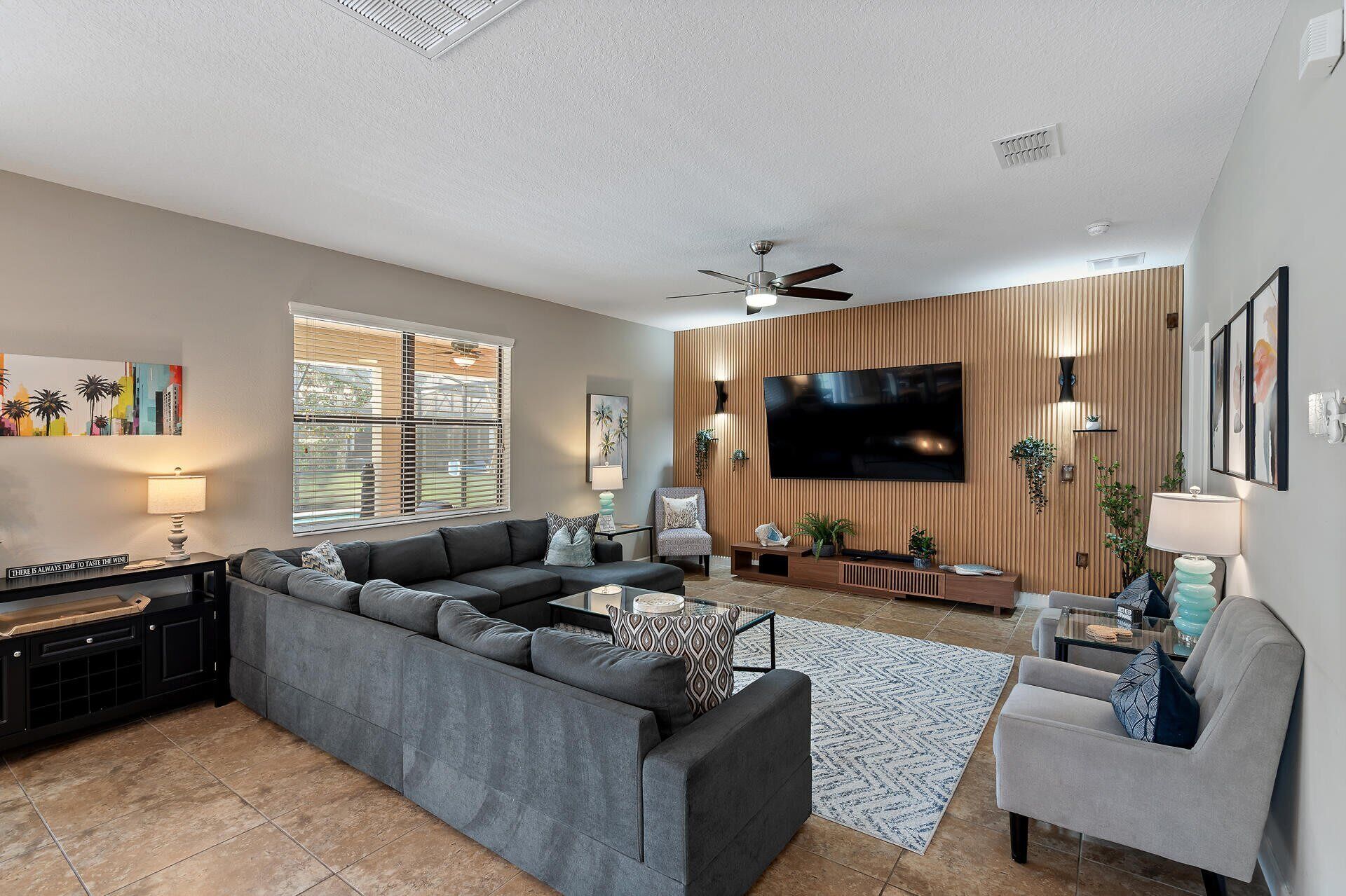 Een moderne woonkamer met een grijze modulaire bank, een flatscreen televisie aan een houten wand bevestigd, een plafondventilator en een groot raam met jaloezieën.