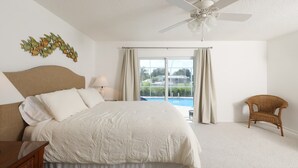 A bedroom with a large bed, a ceiling fan, a window with curtains, a wicker chair, and a view of a swimming pool. - Vgd27305 (Punta Gorda)