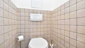 Une petite salle de bain carrelée, équipée de toilettes, d’un porte-rouleau de papier toilette et d’une poignée de chasse d’eau.