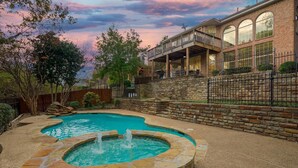 A backyard with a swimming pool and a hot tub, surrounded by a stone wall and a wooden deck. - 5BR Oasis w/ Pool • Steps from Lake Ray Hubbard (Rowlett)