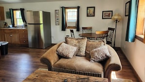 Una sala de estar acogedora con un sofá, una mesa de centro de madera y una zona de comedor con mesa y sillas. Se ve una cocina con refrigerador y armarios al fondo.