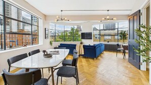 A modern living room with a large window, a blue sofa, a dining table with chairs, and a plant. - Spacious Two-Bedroom Haven by Tech City (Greater London)