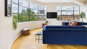 A modern living room with a curved ceiling, large windows offering city views, a blue sofa, and a flat-screen TV. - Spacious Two-Bedroom Haven by Tech City (Greater London)