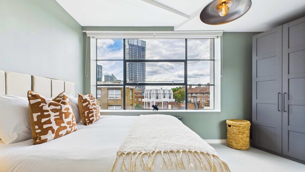 A modern bedroom with a large bed, a view of the city through a large window, and a ceiling light fixture. - Spacious Two-Bedroom Haven by Tech City (Greater London)