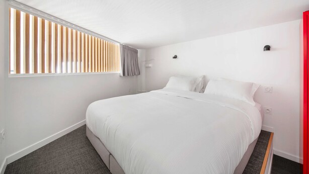 A modern hotel room with a large bed, a window with blinds, a red accent wall, and a small bedside table. - Luxury Apartment 4br/8p - Notre Dame / Bastille (Paris)