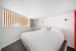 Ein modernes Hotelzimmer mit einem großen Bett, einem Fenster mit Jalousien, einer roten Akzentwand und einem kleinen Nachttisch.