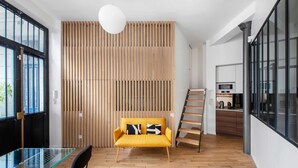 A modern living space with a wooden slatted wall, a yellow sofa with black and white pillows, a glass table with chairs, and a kitchen area with a stove and cabinets. - Luxury Apartment 4br/8p - Notre Dame / Bastille (Paris)