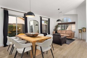 Een moderne woonkamer met een houten eettafel, witte stoelen, een bruine bank en een groot raam.