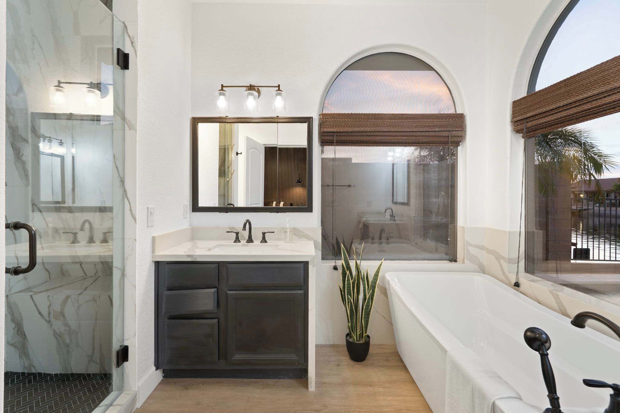 A bathroom with a marble wall, a large mirror, a sink, a bathtub, and a window with blinds.