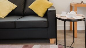 Una sala de estar moderna con un sofá de cuero negro, dos almohadas amarillas y una mesita redonda con una tetera blanca y tazas.