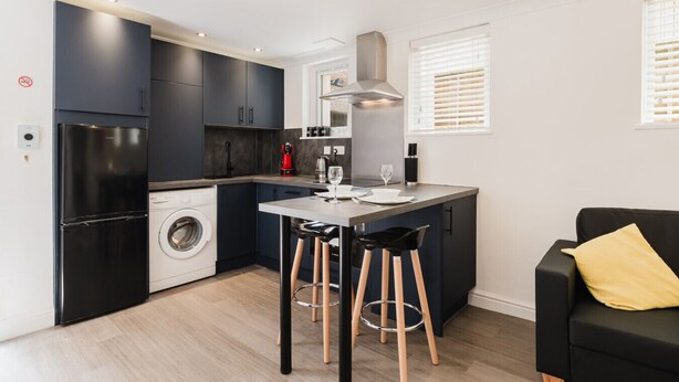 Cocina compacta con armarios oscuros, lavadora, zona de comedor con taburetes y sofá negro con un cojín amarillo.