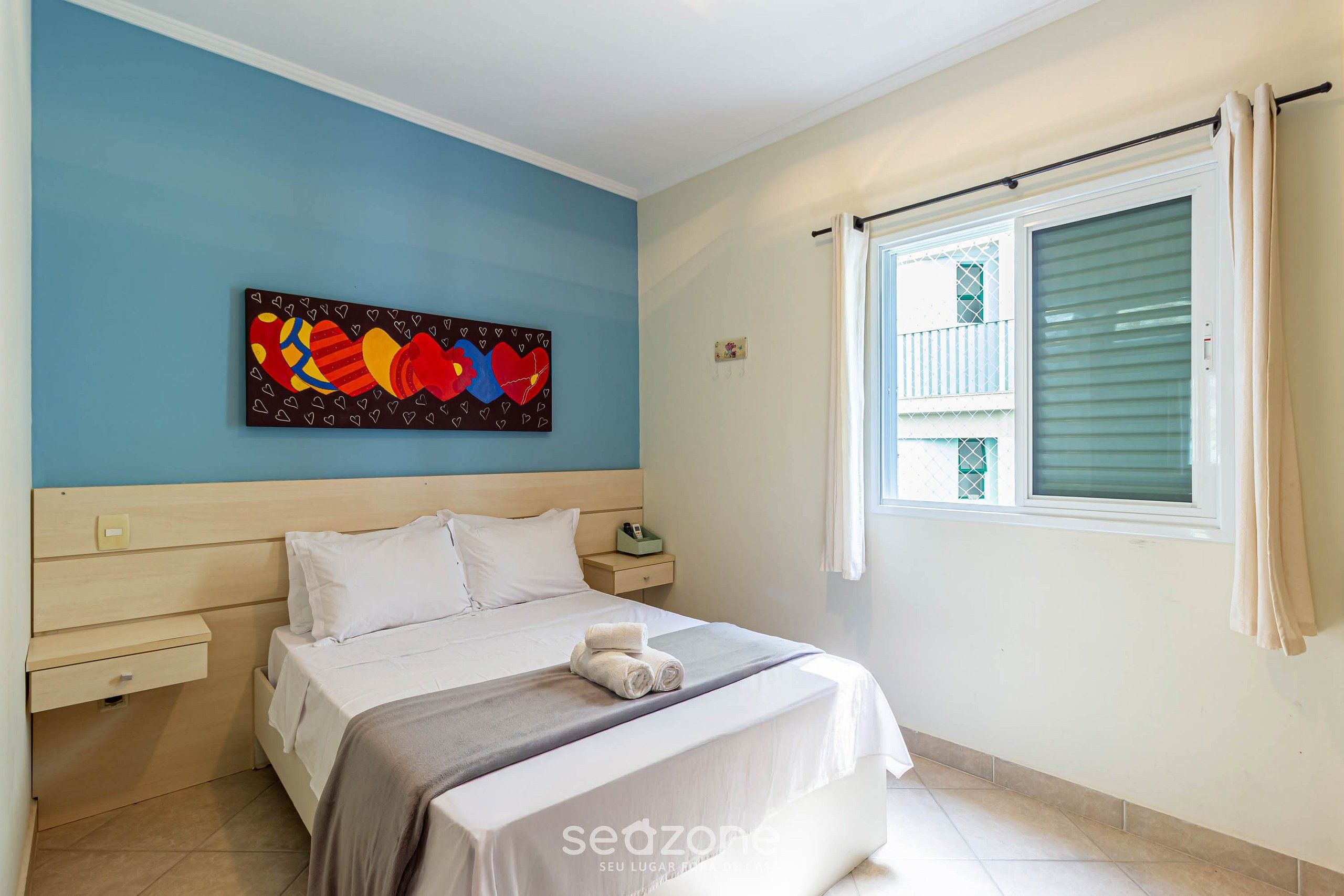 A bedroom with a bed, a nightstand, a wall art piece, a window with curtains, and a tiled floor.