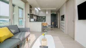 A modern living room with a sofa, a coffee table with a remote control, a kitchen area in the background, and a TV mounted on the wall. - Urbanstay Incheon Songdo (Incheon)