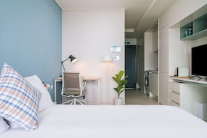 A hotel room with a bed, desk, chair, lamp, potted plant, and a TV.