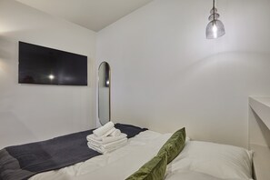 Ein ordentlich bezogenes Bett mit einem gefalteten Handtuch, einem Nachttischspiegel und einem wandmontierten Flachbildfernseher.
