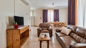 A living room with a brown leather sofa, a wooden coffee table, a TV on a wooden stand, and a dining area with a table and chairs. - Villa Sardinella-5 Bedroom Villa With Private Pool (Mellieha)