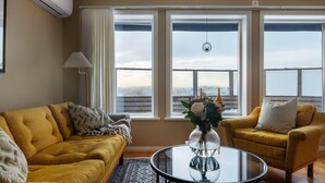 Een woonkamer met een gele bank, een ronde glazen salontafel en een gele fauteuil met een wit kussen. Er zijn drie grote ramen met witte gordijnen, en zicht op een besneeerd landschap.