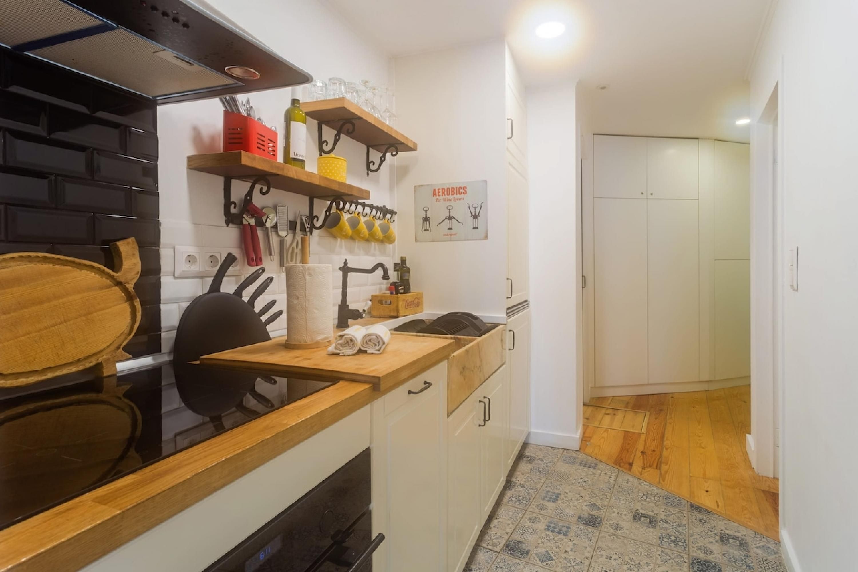 Une cuisine compacte avec un poêle noir, un plan de travail en bois et des placards blancs. On y trouve des étagères remplies d’ustensiles de cuisine et une affiche encadrée accrochée au mur.