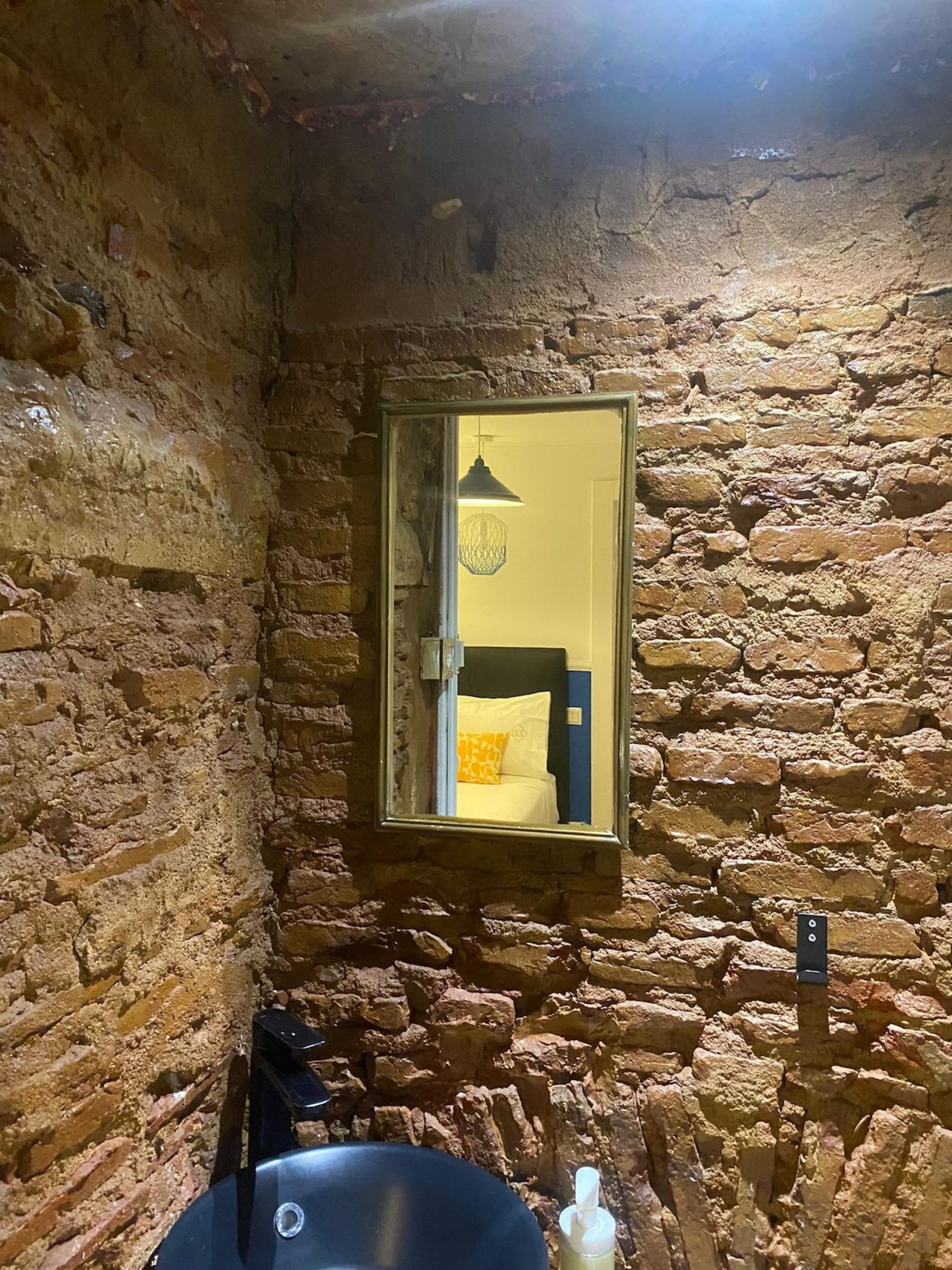 A rustic stone-walled bathroom with a small mirror, a sink, and a hanging light fixture.